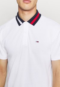 Bílá polo košile s límcem v námořnické modré a červené pruhy. Obsahuje malé vyšitě logo na prsou. Krátké rukávy a standardní střih.