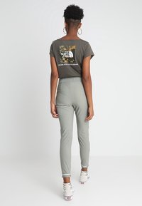 Šedé bavlněné tričko s maskáčovým logem na zadní straně, v kombinaci s světlými zelenými upnutými kalhotami a bílými teniskami. Ohebné náušnice viditelné.