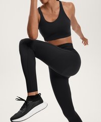 Črn športni modrček in visoke legice iz raztegljive tkanine. Črni in beli športni čevlji z teksturirano zgornjimi deli in belim podplatom.