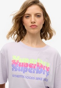 Lavendel oversized t-shirt med et levende flerfarvet grafisk design, der inkluderer teksten "Superdry" og "Authentic Goods Since 2003."