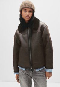 Jonge persoon met een bruine schapenvachtjas, lichtblauw gestreept overhemd, grijze spijkerbroek, beige gebreide muts en zilveren ringen tegen een effen achtergrond.