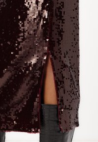Burgunderfarbendes Paillettenkleid mit einem Seitenschlitz, das mit glitzernden, reflektierenden Pailletten und einer glatten Textur versehen ist. Kombiniert mit glänzenden schwarzen Stiefeln.