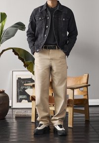 Giubbotto di jeans nero con dettagli strappati, abbinato a pantaloni beige. T-shirt nera sotto e sneakers bianco e nero. Ambiente interno con piante.