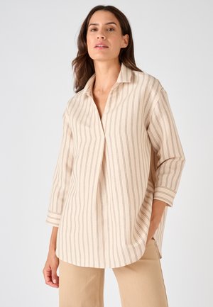 Femme portant une chemise beige ample à fines rayures verticales et un pantalon beige, posant devant un fond gris clair uni.