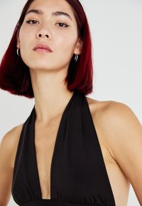 Črna halter obleka z globokim V-izrezom, izdelana iz gladkega materiala in z nabranim pasom. Model prikazuje minimalne dodatke in raven stil frizure.