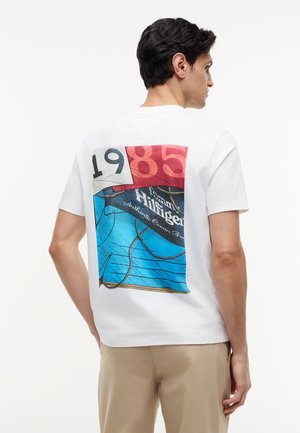 Mann trägt weißes T-Shirt mit farbigem Grafikmotiv und dem Text "1985" auf dem Rücken, kombiniert mit beigen Hosen, seitlich stehend vor weißem Hintergrund.