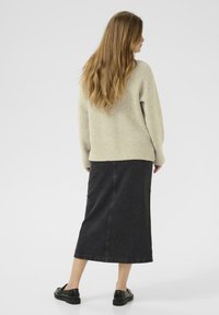 Woman with long hair wearing a beige sweater, black midi denim skirt, and black loafers standing facing away on a plain light background.