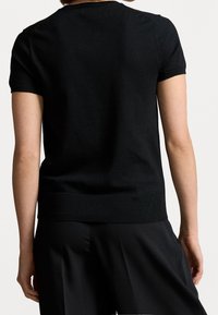 Černý pletený top s krátkým rukávem a žebrovaným výstřihem a dolním lemem. Textura se zdá být hladká, přiléhavá v pase a dobře ladí s černými kalhotami.