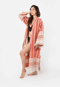 Črtasti kimono v koralni in kremni barvi, z odprtim sprednjim delom, širokimi rokavi in frufrujem na robu. Nositi ga je mogoče čez črne kopalke, minimalistična zasnova.