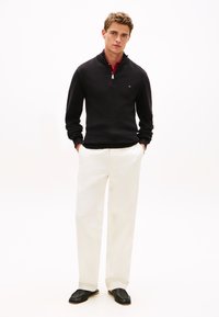 Μαύρο υφασμάτινο πουλόβερ με φερμουάρ, λευκό παντελόνι φαρδιάς γραμμής και μαύρα υφαντά μοκασίνια. Το σύνολο αντιπαραθέτει και ισορροπεί σκούρα και ανοιχτά χρώματα.