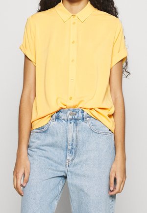 Femme portant une chemise jaune à manches courtes boutonnée, rentrée dans un jean bleu clair taille haute, debout les bras détendus.