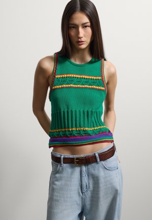 Frau mit langen dunklen Haaren trägt ein grünes gestricktes ärmelloses Oberteil mit mehrfarbigen Mustern und hellblaue Jeans mit braunem Gürtel, steht neutral.