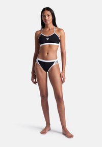 Svart bikinitopp med en bralette överdel och lågsittande underdelar, båda med vit kant. Materialet verkar vara slätt, med minimal mönsteraccent.