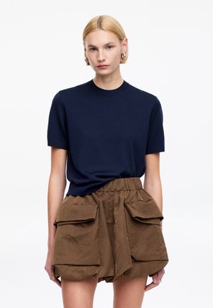 Blondynka w granatowej bluzce z krótkim rękawem i wysokich brązowych szortach z dużymi przednimi kieszeniami na białym tle.