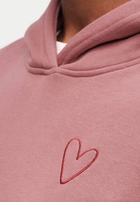 Nahaufnahme eines pinkfarbenen Kapuzenpullovers mit einem einfachen roten Herz, das auf der Brust in der Nähe des Kragens gestickt ist.