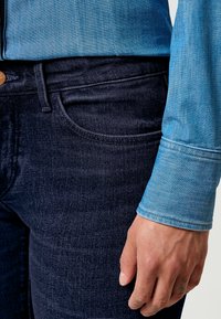 Jeans de mezclilla en azul oscuro con una textura suave, que presentan un diseño clásico de cinco bolsillos y un cierre con botón en la parte superior. Camisa de mezclilla en azul claro visible.