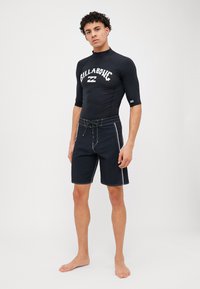 Fekete rash guard rövid ujjakkal, fehér "BILLABONG" logóval, sötét színű fürdőnadrággal párosítva, amelyen fehér varrás és húzózsinór található.