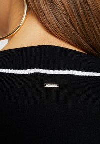 Schwarzer Strickpullover mit einem weißen Streifen entlang des hinteren Halsausschnitts, mit einem kleinen silbernen Markentag in der Mitte befestigt.