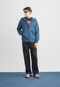 Sweat à capuche zippé bleu avec capuche, associé à un pantalon noir avec des coutures rouges. Le mannequin porte des lunettes de soleil et un t-shirt noir graphique.