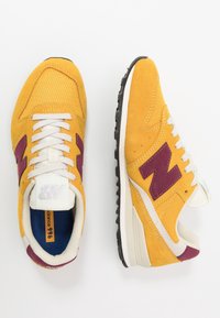 Gula sportskor med ovandel i mocka och mesh, med en vinröd logotyp, vita skosnören och en svart gummisula.