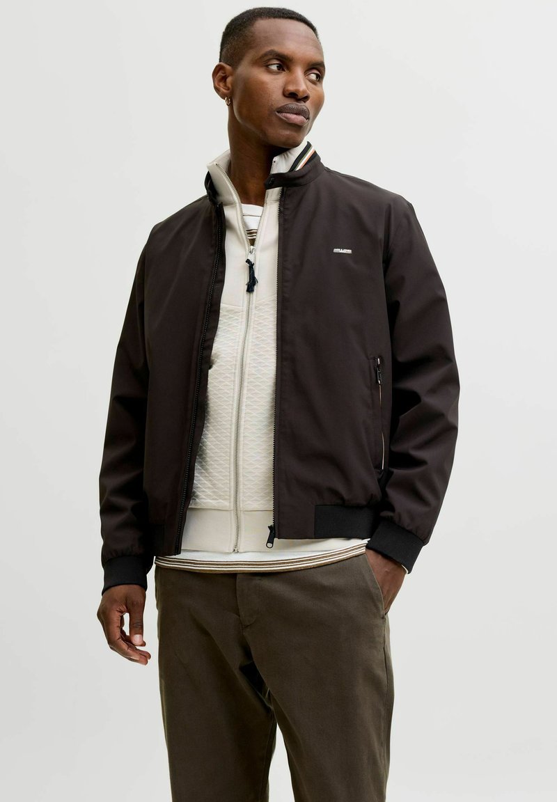 Νέος άνδρας με μαύρο μπουφάν bomber, λευκό πουλόβερ με φερμουάρ και καφέ παντελόνι, στέκεται με το ένα χέρι στην τσέπη μπροστά σε μονόχρωμο φόντο.