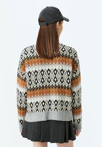 Gestrickter Pullover mit einem geometrischen Muster in Orange, Schwarz und Weiß, mit einem grauen gerippten Saum; über einem plissierten dunklen Rock und einer Mütze getragen.