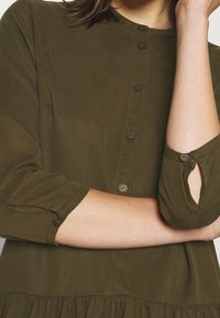 Kobieta ubrana w oliwkową sukienkę z guzikami, z rękawami o długości 3/4 i lekko falbaniastego dołu, z rękami skrzyżowanymi na talii.