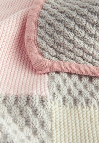 Textured knitted blanket in light pink, gray, and cream, featuring distinct stitch patterns and contrasting edges. Cozy and soft texture.