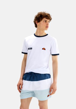Junger Mann trägt weißes T-Shirt mit marineblauem Besatz und Logo, kombiniert mit blau-weißen Colorblock-Shorts mit Logo, steht vor einem einfarbigen Hintergrund.
