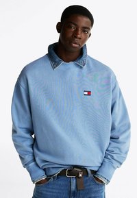 Jasnoniebieski sweterek z dżinsowym kołnierzem, ściągaczami przy rękawach i małą naszywką z logo w kolorach czerwonym, białym i niebieskim. Noszony z niebieskimi dżinsami i czarnym paskiem.