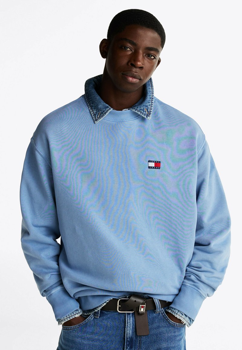 Jasnoniebieski sweterek z dżinsowym kołnierzem, ściągaczami przy rękawach i małą naszywką z logo w kolorach czerwonym, białym i niebieskim. Noszony z niebieskimi dżinsami i czarnym paskiem.