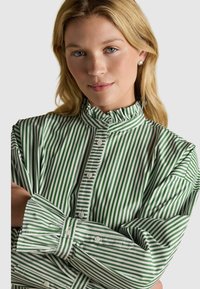 Cămașă cu dungi verzi și albe, cu guler volan, mâneci lungi și detalii la manșete. Material moale cu o textură netedă.