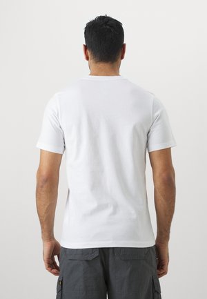 Mann mit kurzen dunklen Haaren, der ein schlichtes weißes kurzärmeliges T-Shirt und dunkelgraue Cargoshorts trägt und mit dem Rücken zur Kamera steht.
