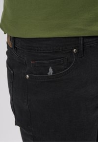 Jeans de mezclilla negra con un bolsillo frontal que presenta un pequeño logo bordado, herrajes plateados y un detalle de costura roja en contraste.