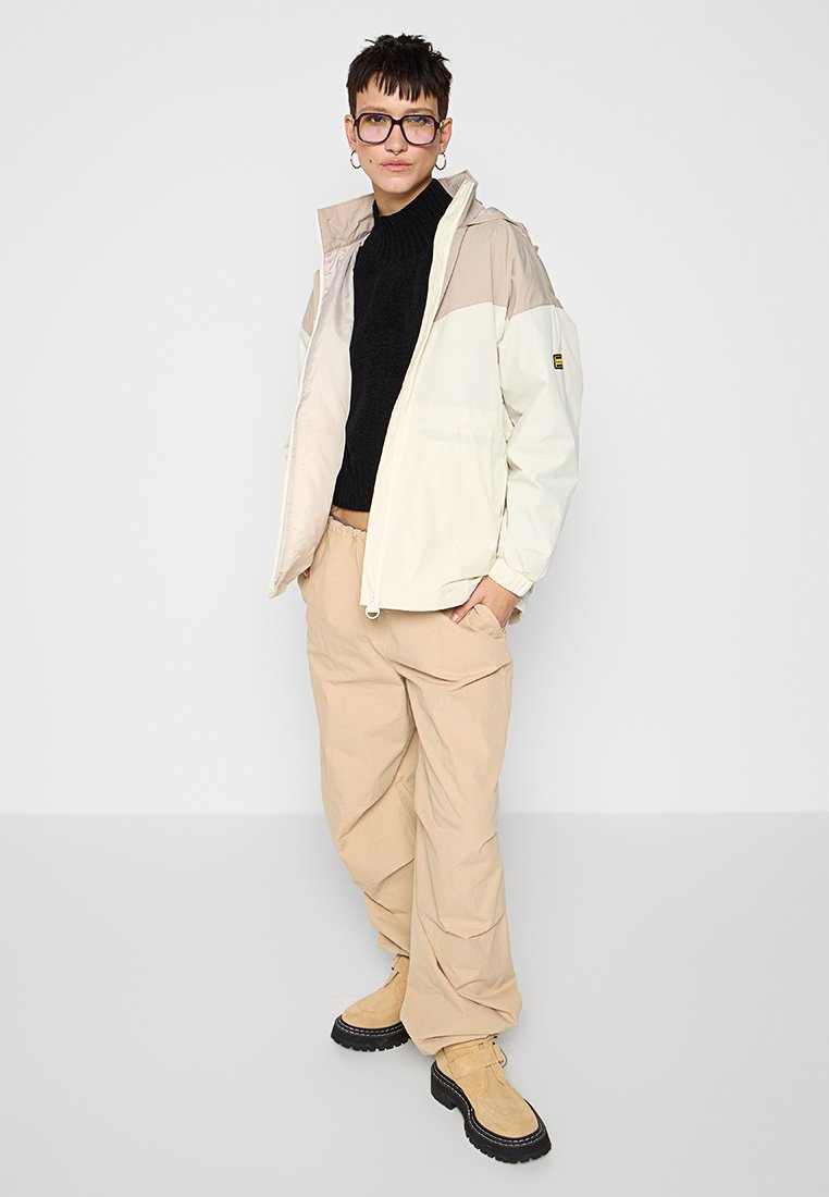 Giacca leggera beige e crema con colletto alto, abbinata a pantaloni larghi beige e stivali beige con lacci; presenta un maglione nero sotto.