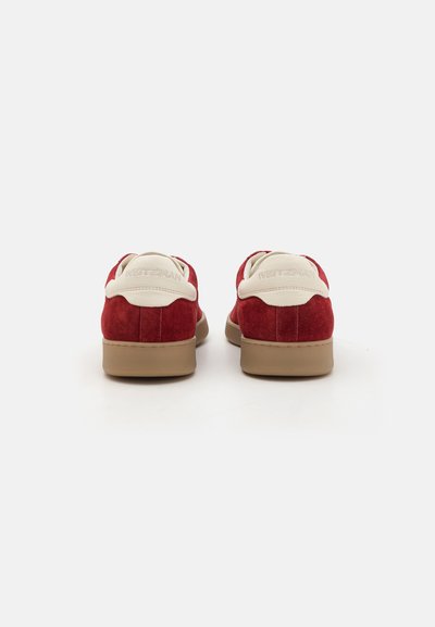 Baskets en daim rouge avec un patch en cuir beige au niveau du talon et une semelle en caoutchouc marron. Présentent un logo en relief sur le talon et une texture lisse.