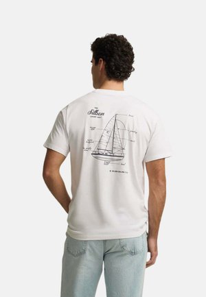 Hombre vistiendo una camiseta blanca con un diagrama y etiquetas de un velero en negro impresas en la espalda, combinado con vaqueros azul claro.