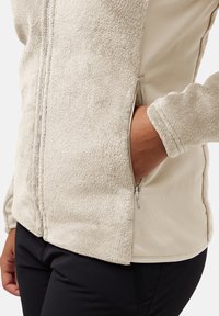 Giacca in pile beige con una texture morbida, dotata di tasche laterali con zip e design aderente. Le maniche hanno una vestibilità attillata e un tessuto liscio.