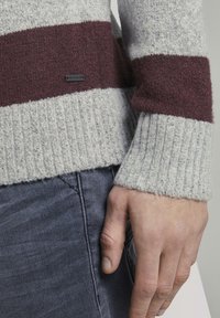 Main d'homme posée à côté d'un jean bleu, portant un pull tricoté avec de larges rayures horizontales grises et bordeaux.