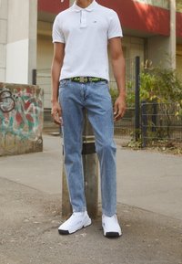 Vit polo-shirt med krage, ljusblå raka jeans och vita slipsneakers med svarta detaljer, som står mot en betongbarriär.