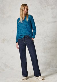 Blusa blu a fantasia con design a rombi, maniche lunghe e un sottile scollo a V; abbinata a jeans dritti blu scuro e sneaker beige.