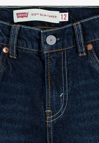 Tmavomodré denimové nohavice Levi's 512 Slim Taper majú žlté šitie, kovový gombík s logom a bielu etiketu s červeným textom. Veľkosť 12.