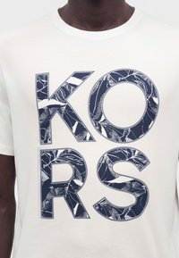 Κοντινή λήψη ενός λευκού μπλουζάκι με μεγάλα σκούρα μπλε γράμματα "KORS" γεμάτα με λευκό και σκούρο μπλε φύλλωμα.