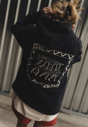 Osoba o blond włosach związuje kucyk, ubrana w czarną bluzę z kapturem z białym tropikalnym kwiatem i grafiką deski surfingowej na plecach.