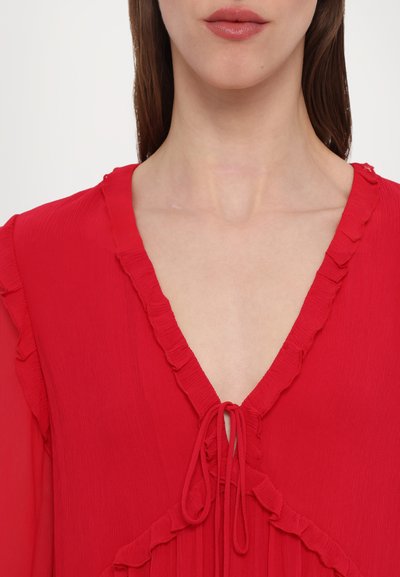 Blouse rouge avec un col en V froncé et un détail de nœud, fabriquée en tissu léger avec une finition texturée.