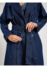 Trench coat de mezclilla en azul oscuro, con un frente de doble botonadura, amplios solapas, cinturón en la cintura y acentos de botón en las mangas.