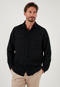 Cămașă neagră cu mânecă lungă, fabricată dintr-un material moale, având două buzunare pe piept și închidere cu nasturi. Designul dispune de un guler clasic și tiv drept.