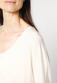 Světle béžový top s širokým výstřihem a krátkými rukávy, s měkkou a hladkou texturou. Jemný žebrovaný detail podél látky.