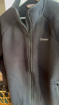 Donkergrijze fleecejas met volledige ritssluiting aan de voorkant, zijzakken en Berghaus-logo op de borst, hangend aan een houten hanger.