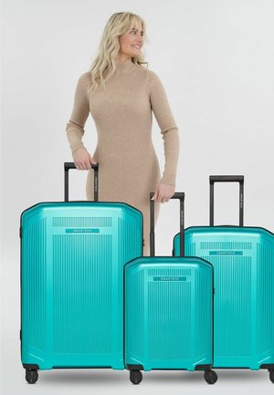 Vrouw in beige jurk die handgrepen vasthoudt van drie turquoise koffers met een harde schaal in verschillende maten op wieltjes.